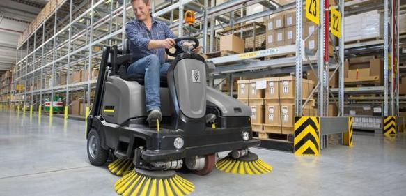 Operator na samojezdnej maszynie czyszczącej z żółtymi szczotkami, w magazynie z regałami wypełnionymi kartonami.
