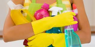 Osoba w żółtych rękawicach ochronnych trzymająca w ramionach różnorodne środki czystości w plastikowych butelkach, widoczne fragmenty fartucha.