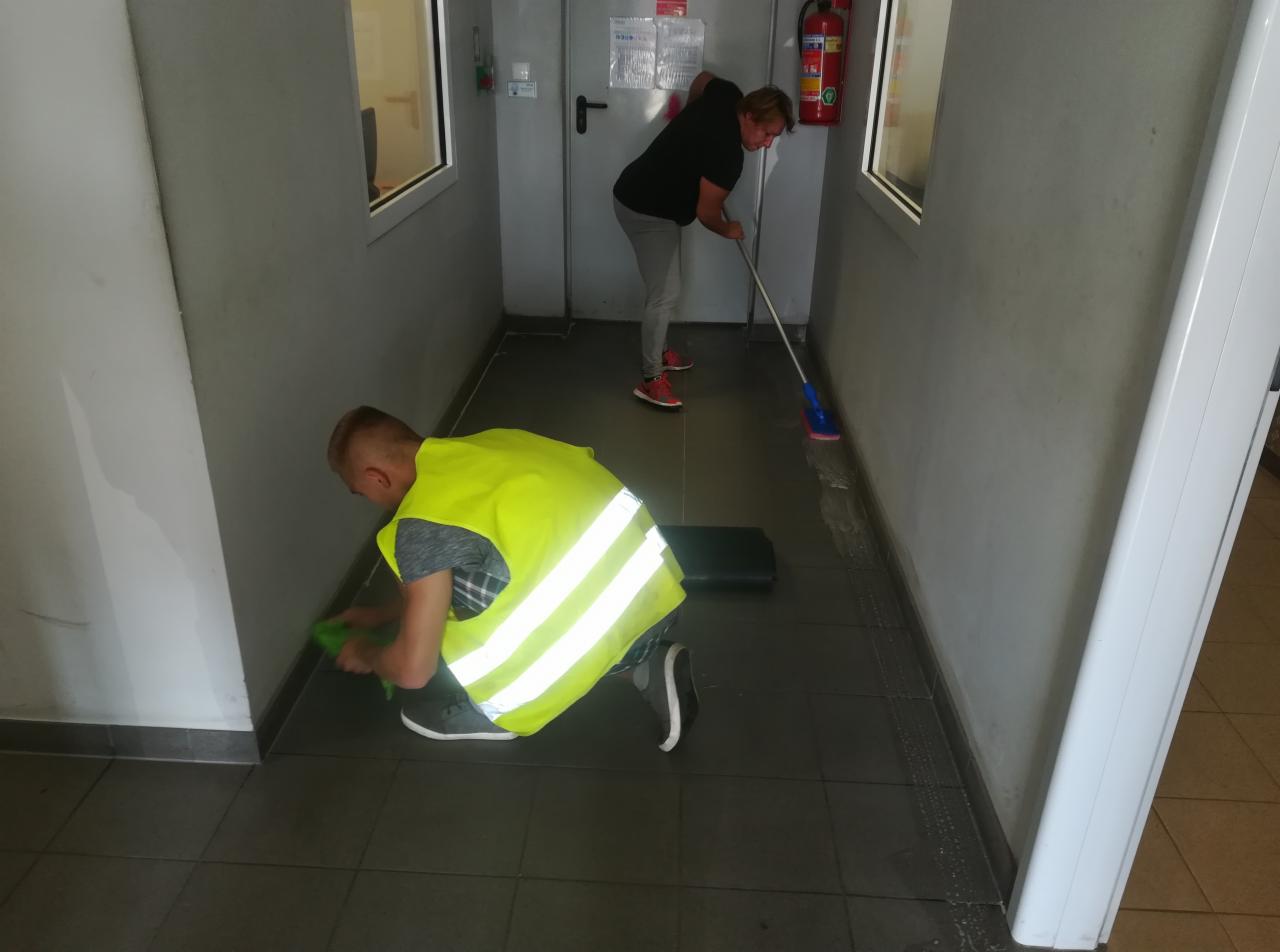 Dwóch pracowników sprzątających korytarz biurowy: jeden czyści narożnik ściany zieloną gąbką, drugi myje podłogę mopem, widoczny ślad mokrej podłogi.