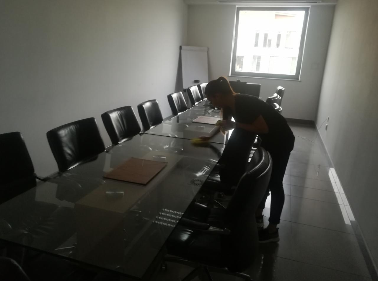 Pracownica w czarnej koszulce i dżinsach przeciera wilgotną ściereczką długi, szklany stół konferencyjny w biurze, na którym leżą teczki, a wokół stoją czarne, skórzane krzesła.