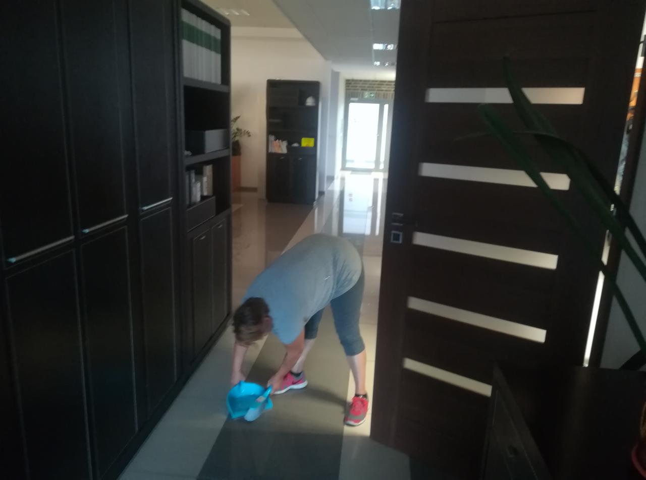 Kobieta w szarym T-shircie i legginsach, pochylona, myje podłogę w biurze za pomocą niebieskiego wiadra i mopa, widoczne ciemne szafy i drzwi.