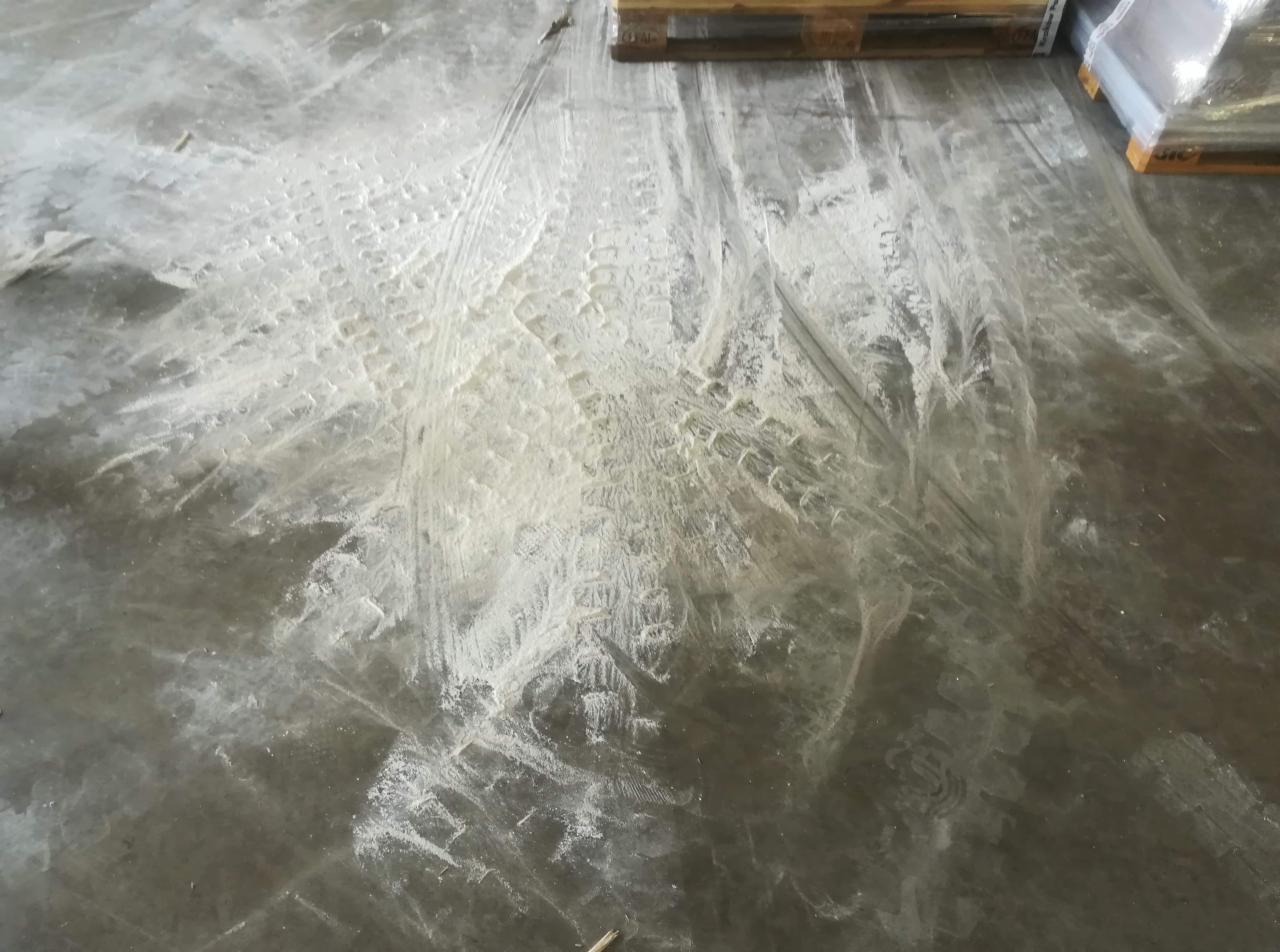 Betonowa podłoga po remoncie z widocznymi śladami opon i resztkami białego pyłu budowlanego, w tle palety z materiałami budowlanymi.