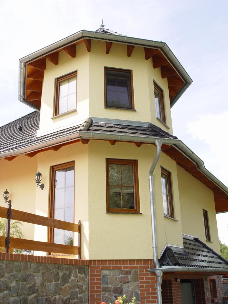 Dwupiętrowy dom z oknami PCV w drewnianych ramach, z widocznymi rynnami i fragmentem kamienno-ceglanego ogrodzenia.