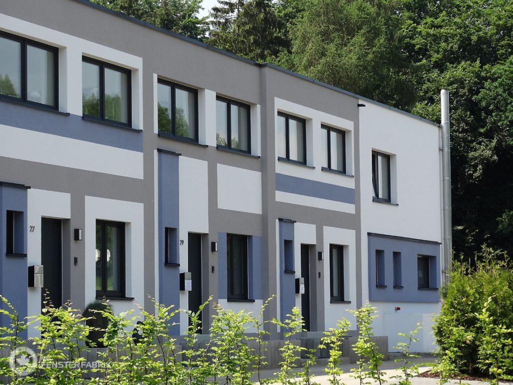 Elewacja szeregówki z czarnymi oknami, pomalowana w geometryczne wzory w odcieniach szarości i bieli, z zielenią w tle i na pierwszym planie.