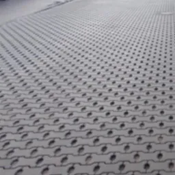 Szara, ażurowa nawierzchnia z kostki betonowej z otworami, ułożona na parkingu, widok z perspektywy chodnika, w tle zaparkowany samochód i budynek.