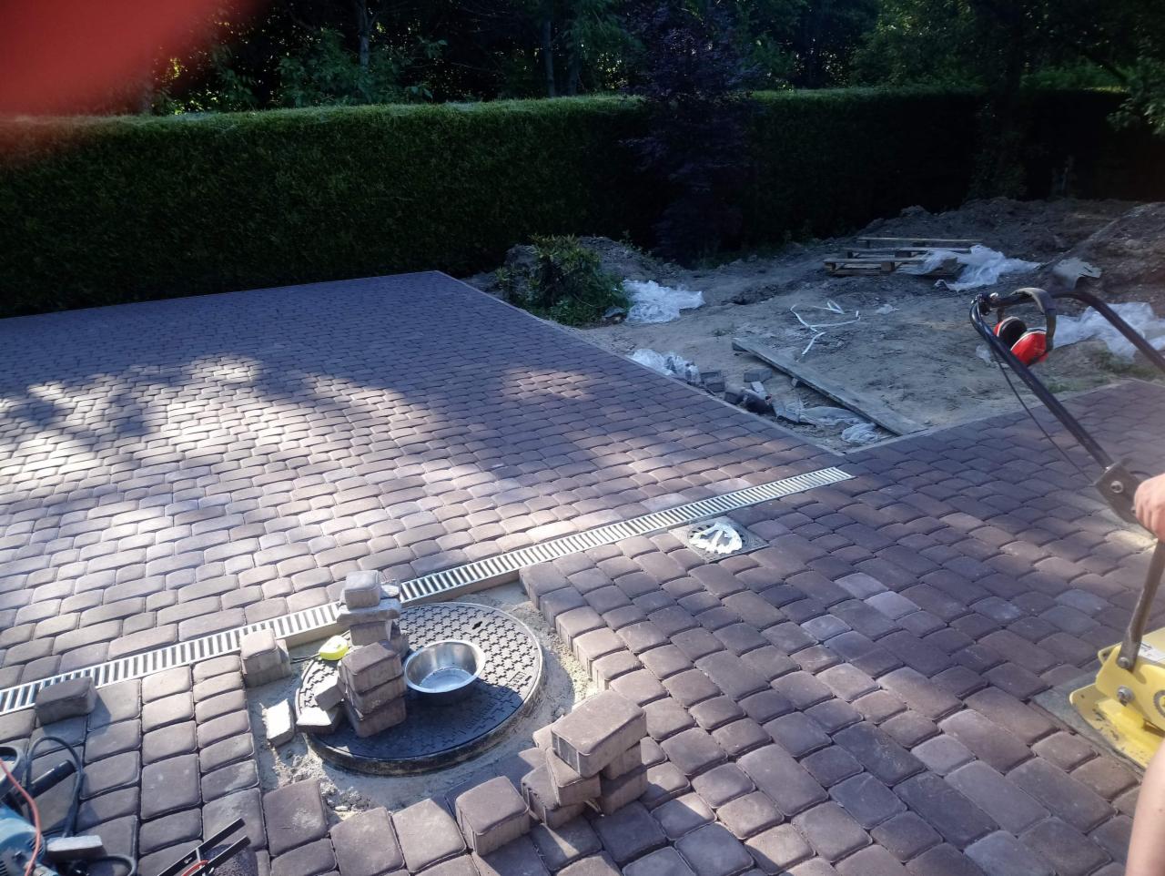 Prace brukarskie w toku: ułożona część nawierzchni z kostki w odcieniach brązu, widoczne korytka odwadniające, studzienka kanalizacyjna z metalową miską i stosem kostek, w tle przygotowany teren...