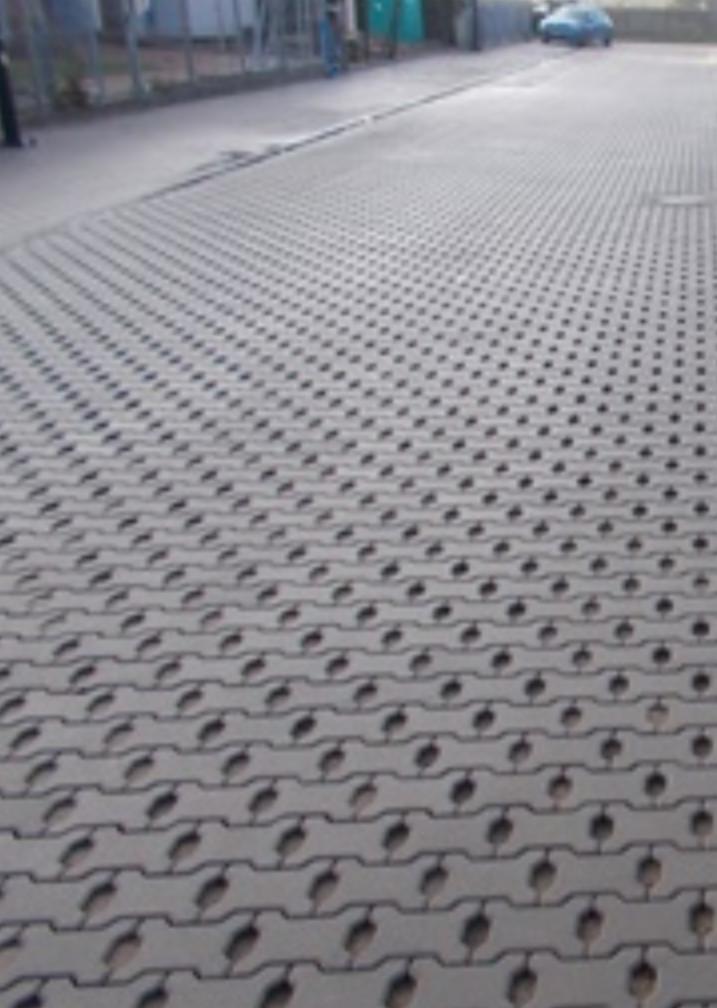 Szara, ażurowa nawierzchnia z kostki betonowej z otworami, ułożona na parkingu, widok z perspektywy chodnika, w tle zaparkowany samochód i budynek.