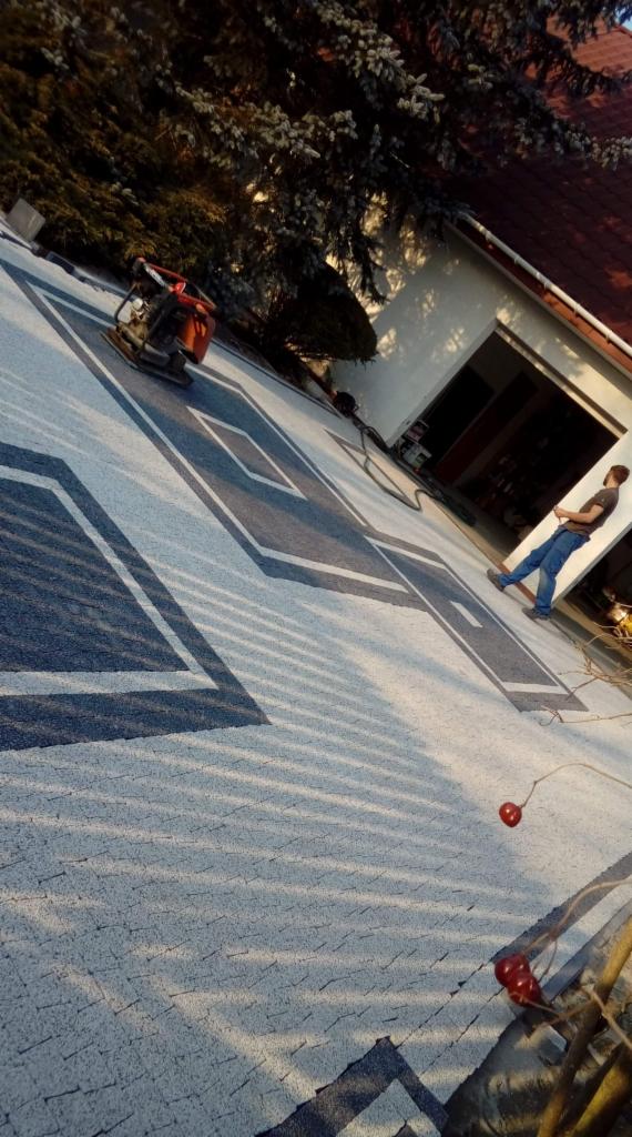 Wzorzysta nawierzchnia z jasnoszarej kostki z ciemnoszarymi geometrycznymi wstawkami, zagęszczarka wibracyjna w rogu, mężczyzna w jeansach i T-shircie stoi obok otwartego garażu, drzewo iglaste w tle.