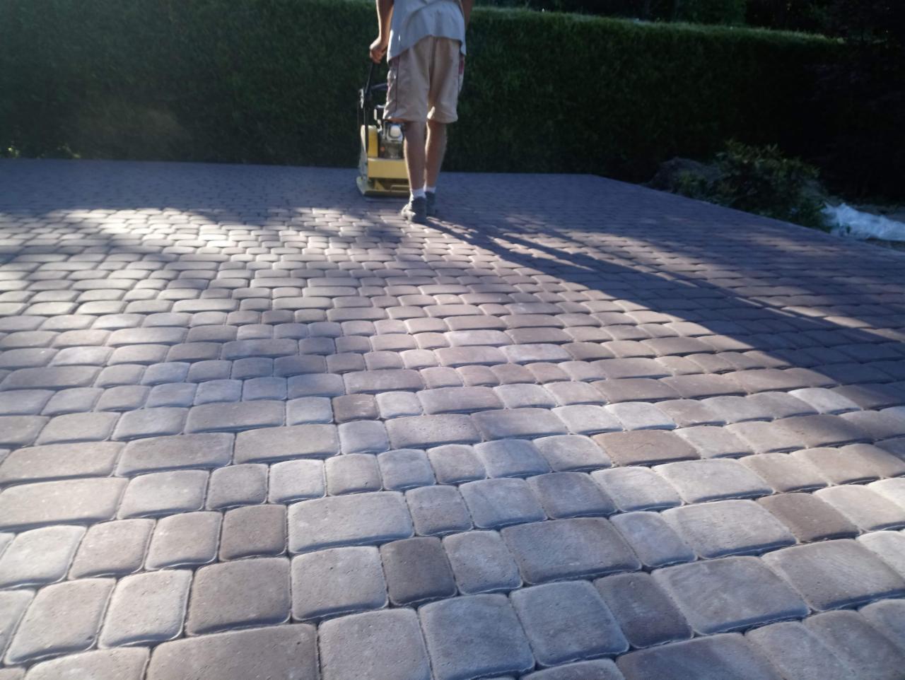 Prace brukarskie: Zagęszczanie nowo ułożonej nawierzchni z kostki w odcieniach szarości i brązu przy użyciu ubijarki wibracyjnej, z widocznym cieniem operatora i maszyny na powierzchni.