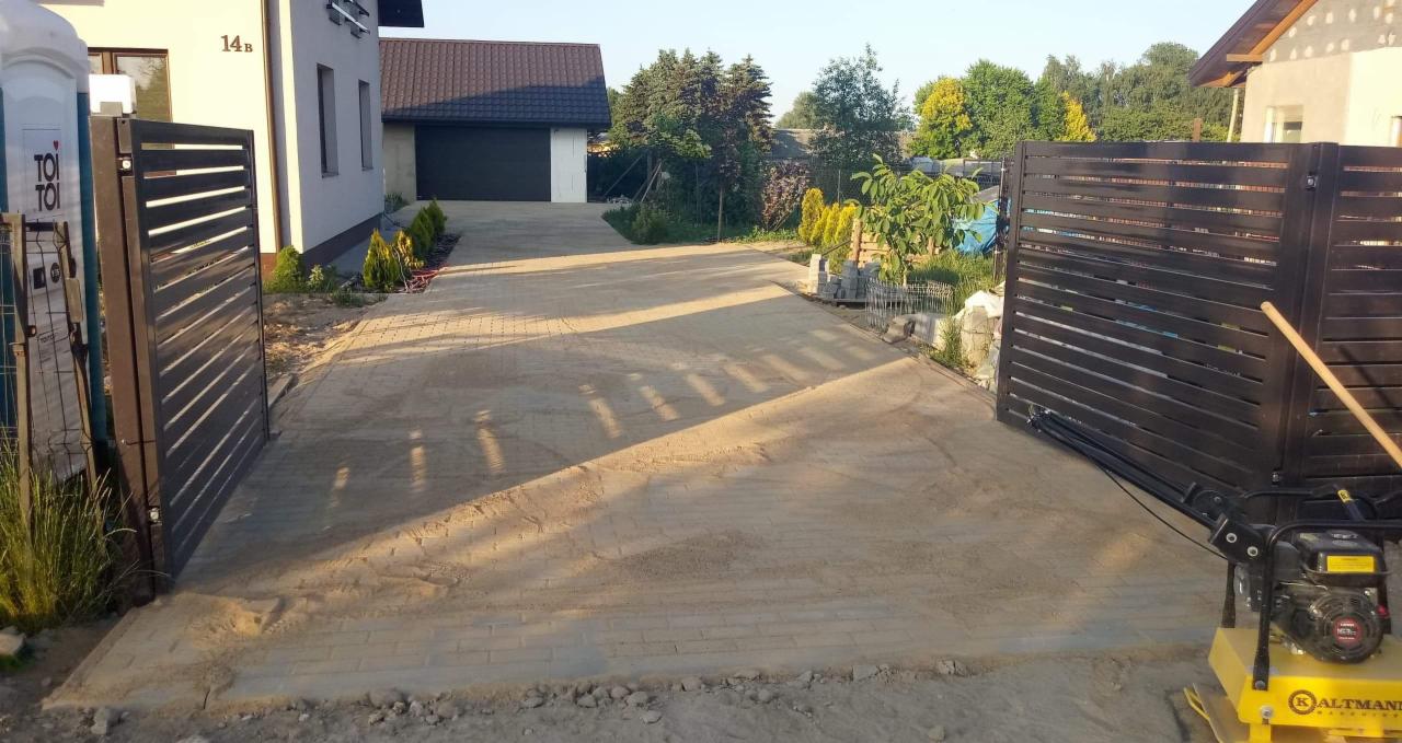 Niedokończona, piaskowa nawierzchnia podjazdu z widoczną ubijarką w tle i metalowymi bramami po bokach, dom z ciemnym dachem w tle.