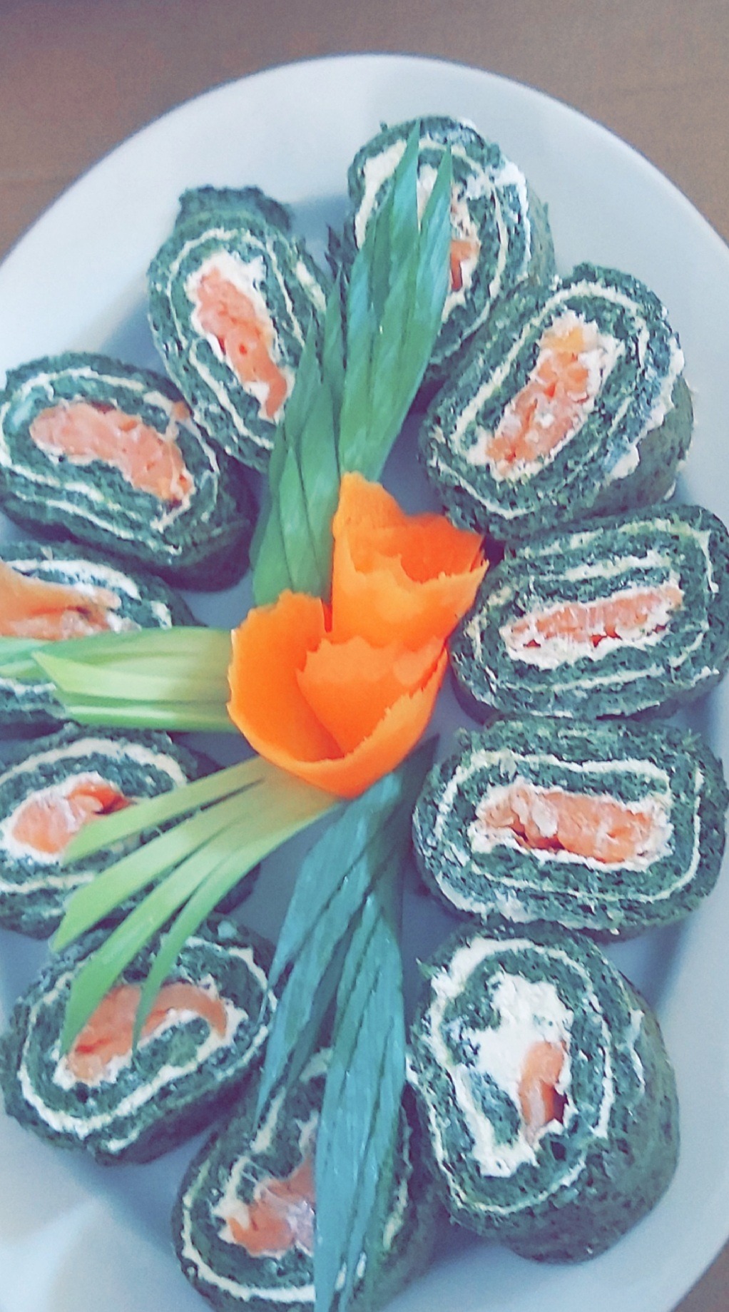 Dekoracyjnie ułożone roladki ze szpinaku z łososiem i kremowym serem, ozdobione marchewkową rozetą i pędami cebuli na białym talerzu.