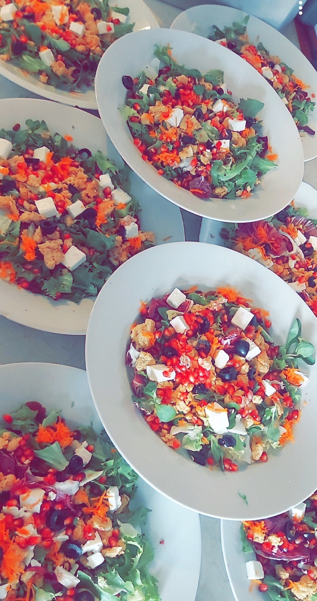 Porcje sałatek z granatem, serem feta, oliwkami i marchewką na białych talerzach, przygotowane do serwowania.