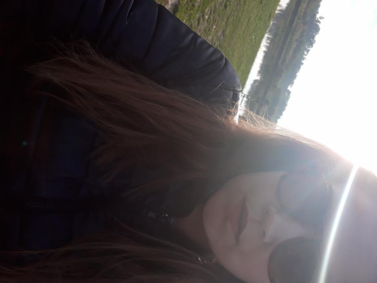 Portret kobiety w okularach przeciwsłonecznych, z długimi brązowymi włosami i w kurtce puchowej, na tle rozmytego krajobrazu z zielenią i wodą, z silnym blaskiem słonecznym.