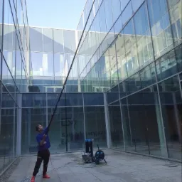 Pracownik w czerwonych butach myje szklaną fasadę budynku biurowego za pomocą długiej lancy i profesjonalnego sprzętu Ionic. Widoczny napis 'Sala edukacyjna'.