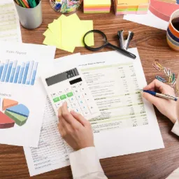 Kobieta analizuje dokumenty finansowe przy biurku z kalkulatorem, długopisem, spinaczami, kubkiem z kawą i raportami sprzedaży z wykresami.