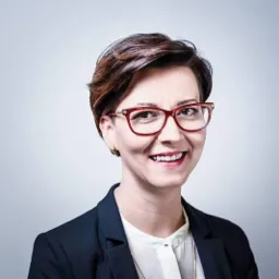 Uśmiechnięta kobieta w okularach z czerwoną oprawką, ubrana w czarną marynarkę i białą koszulę, portret na jasnym tle.