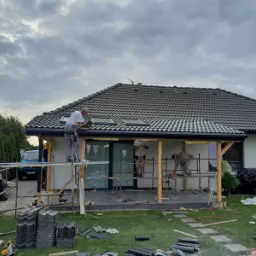 Prace dekarskie przy domu jednorodzinnym: montaż dachówek na dachu z oknami połaciowymi, widoczne rusztowania i stosy dachówek na trawniku.