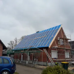 Ceglany dom jednorodzinny z częściowo zdjętym dachem, zabezpieczony niebieską folią i drewnianą konstrukcją, rusztowania i drabina przy ścianie, widoczny samochód i sąsiedni budynek.