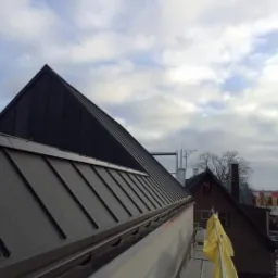 Nowy, ciemny, metalowy dach z blachy trapezowej na tle pochmurnego nieba, widok z rusztowania z żółtą plandeką.