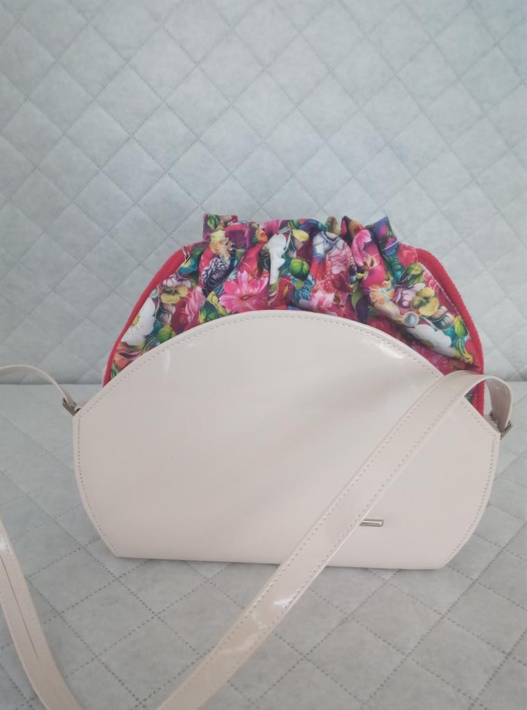 Beżowa lakierowana torebka z materiałową wstawką w kwiatowy wzór, leżąca na pikowanej tkaninie.