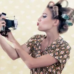 Kobieta w wałkach na włosach robi selfie starodawnym aparatem, usta ułożone w dziubek, wzorzysta bluzka, żółte tło w drobne wzorki.