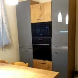 Szara zabudowa kuchenna z piekarnikiem i mikrofalówką, fronty drewnopodobne w górnej i dolnej części, widoczny fragment drewnianego stołu.