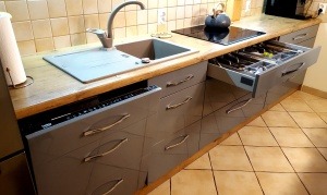 Szare szafki kuchenne z drewnianym blatem, widoczny otwarty szuflada z wkładem na sztućce, bateria kuchenna i płyta indukcyjna na blacie, beżowe płytki na ścianie i podłodze.