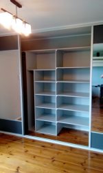 Wnętrze szafy wnękowej z przesuwnymi drzwiami i otwartymi półkami, widok na drewnianą podłogę, oświetlenie sufitowe w tle.