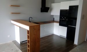 Nowoczesna, jasna kuchnia z białymi szafkami, drewnianym blatem i wyspą z regałem na wino. Czarny okap i sprzęt AGD. Dwie drewniane półki na ścianie. Minimalistyczny design.