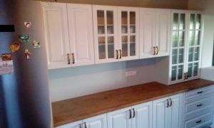 Biała zabudowa kuchenna z drewnianym blatem; górne szafki częściowo przeszklone, dolne z szufladami; widoczny fragment lodówki z magnesami.