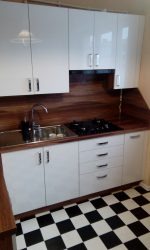 Biała kuchnia z błyszczącymi frontami, brązowym blatem i czarno-białą podłogą w szachownicę. Widoczny zlew ze srebrną baterią i płyta grzewcza. Brązowy panel nad blatem.