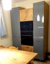 Szara zabudowa kuchenna z piekarnikiem i mikrofalówką, fronty drewnopodobne w górnej i dolnej części, widoczny fragment drewnianego stołu.