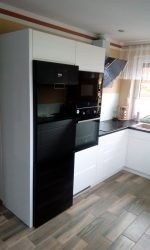 Biała zabudowa kuchenna z czarnymi piekarnikami i mikrofalówką w rogu kuchni, widoczne jasne panele podłogowe i fragment okna z zasłoną.