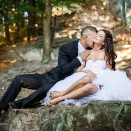 Para młoda w plenerze: pan młody w czarnym garniturze całuje pannę młodą w sukni ślubnej z gorsetem, siedzącą na korzeniach drzew w lesie, z delikatnym uśmiechem i zamkniętymi oczami.