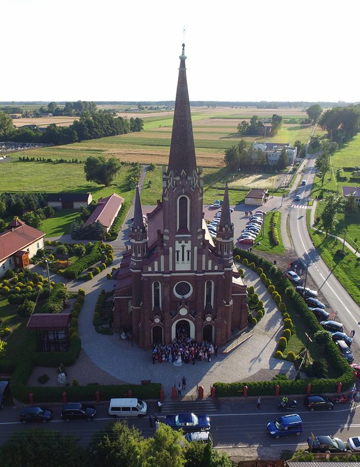 Widok z lotu ptaka na ceglany kościół z wysoką wieżą, przed którym zgromadziła się grupa ludzi, prawdopodobnie po ceremonii ślubnej, z zaparkowanymi samochodami wzdłuż drogi i zielonym otoczeniem.