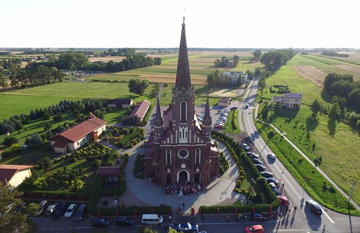 Panorama z lotu ptaka na ceglany kościół z wysoką wieżą, otoczony zielenią, z grupą ludzi przed wejściem, prawdopodobnie po ceremonii ślubnej, i zaparkowanymi samochodami wzdłuż drogi.