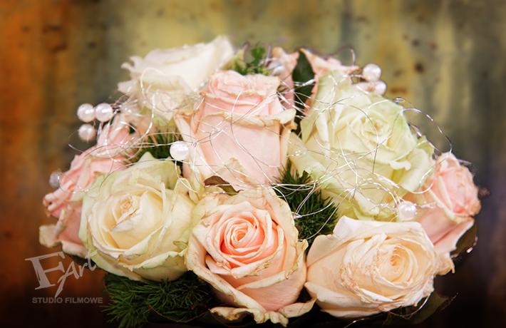 Bukiet ślubny z bladoróżowych i kremowych róż, ozdobiony drobnymi perłami i srebrnym drucikiem, na rozmytym tle w odcieniach brązu i zieleni; widoczne logo studia filmowego w lewym dolnym rogu.