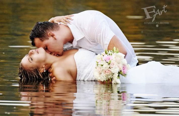 Para młoda w wodzie, mężczyzna całuje kobietę w szyję, bukiet różowych i białych kwiatów, odbicie w wodzie, logo studia filmowego w prawym górnym rogu.