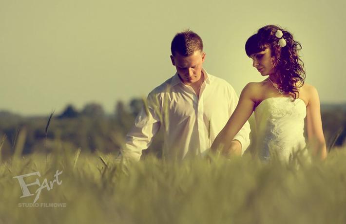 Para młoda w polu pszenicy, panna młoda w sukni bez ramiączek z kwiatami we włosach, pan młody w białej koszuli, delikatne oświetlenie, logo studia filmowego w lewym dolnym rogu.