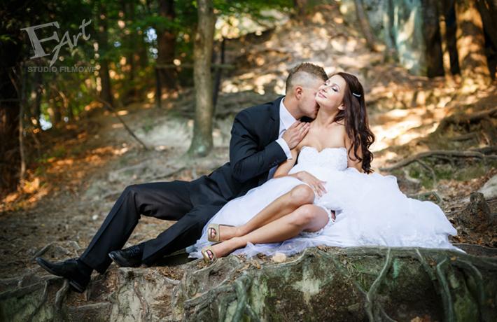 Para młoda w plenerze: pan młody w czarnym garniturze całuje pannę młodą w sukni ślubnej z gorsetem, siedzącą na korzeniach drzew w lesie, z delikatnym uśmiechem i zamkniętymi oczami.