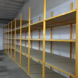Długi rząd pustych, żółtych metalowych regałów magazynowych z płytami wiórowymi na półkach, widoczny w hali magazynowej z betonową podłogą i białymi ścianami.
