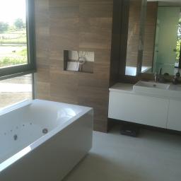 FHU. PROFIKOM Mariusz Jankowicz - Nowoczesna łazienka z dużą wanną z hydromasażem przy oknie z widokiem na zieleń, drewnopodobne płytki na ścianie z wnęką i białymi kablami, oraz biała szafka z umywalką, odbicie osoby robiącej...