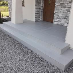 FHU. PROFIKOM Mariusz Jankowicz - Wykończone schody wejściowe do domu, pokryte szarymi płytkami ceramicznymi, otoczone szarym żwirem, z widoczną częścią elewacji z cegły i drewnianymi drzwiami.