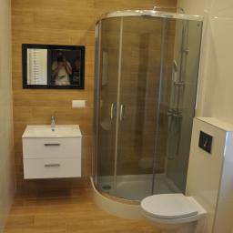 FHU. PROFIKOM Mariusz Jankowicz - Jasna łazienka z kabiną prysznicową narożną, wiszącą szafką z umywalką i drewnopodobnymi płytkami na ścianach, z lustrem odbijającym osoby robiące zdjęcie.