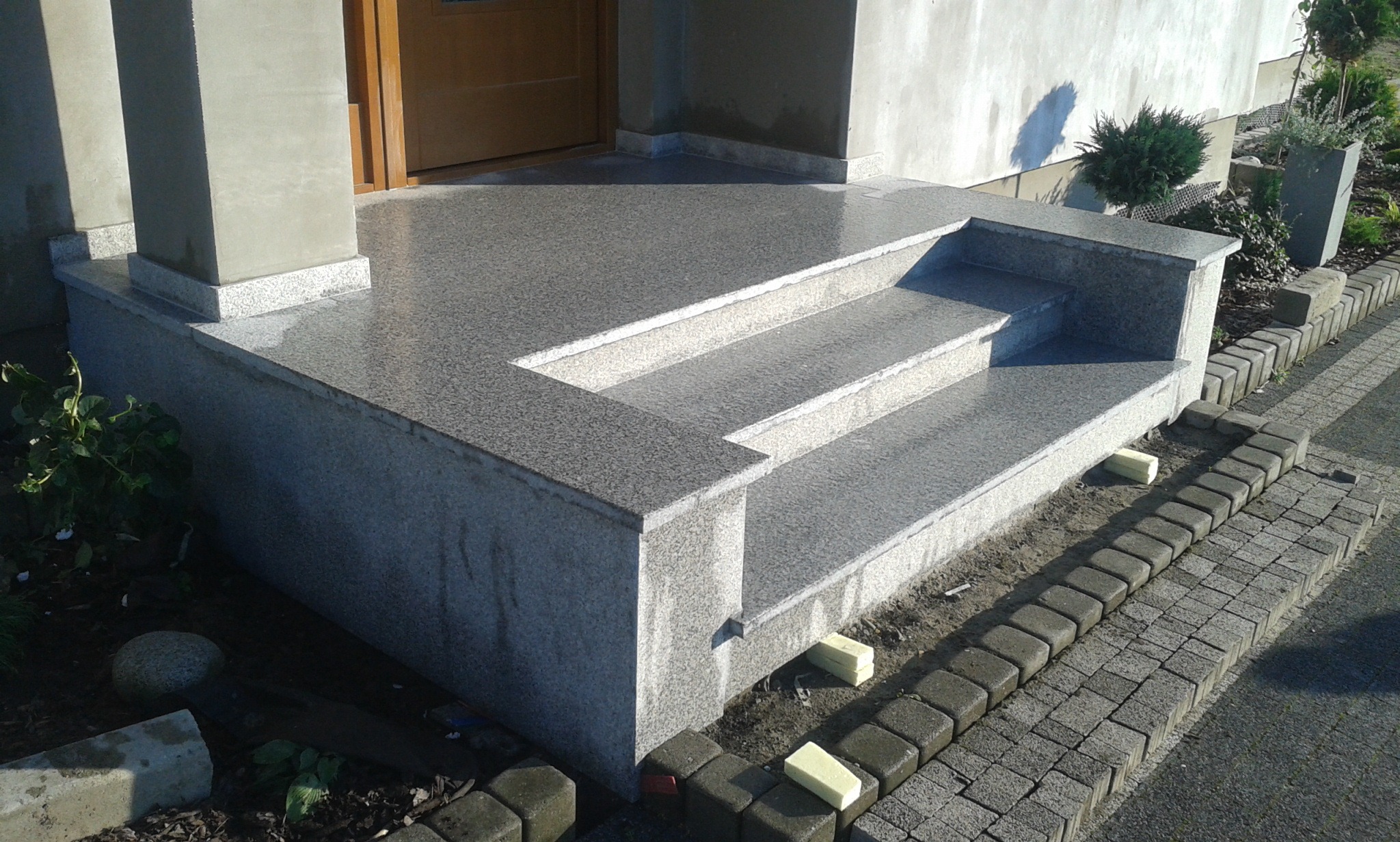 Wykończone granitowe schody wejściowe do domu w Bochni, z widocznymi elementami kostki brukowej i zielenią w tle.