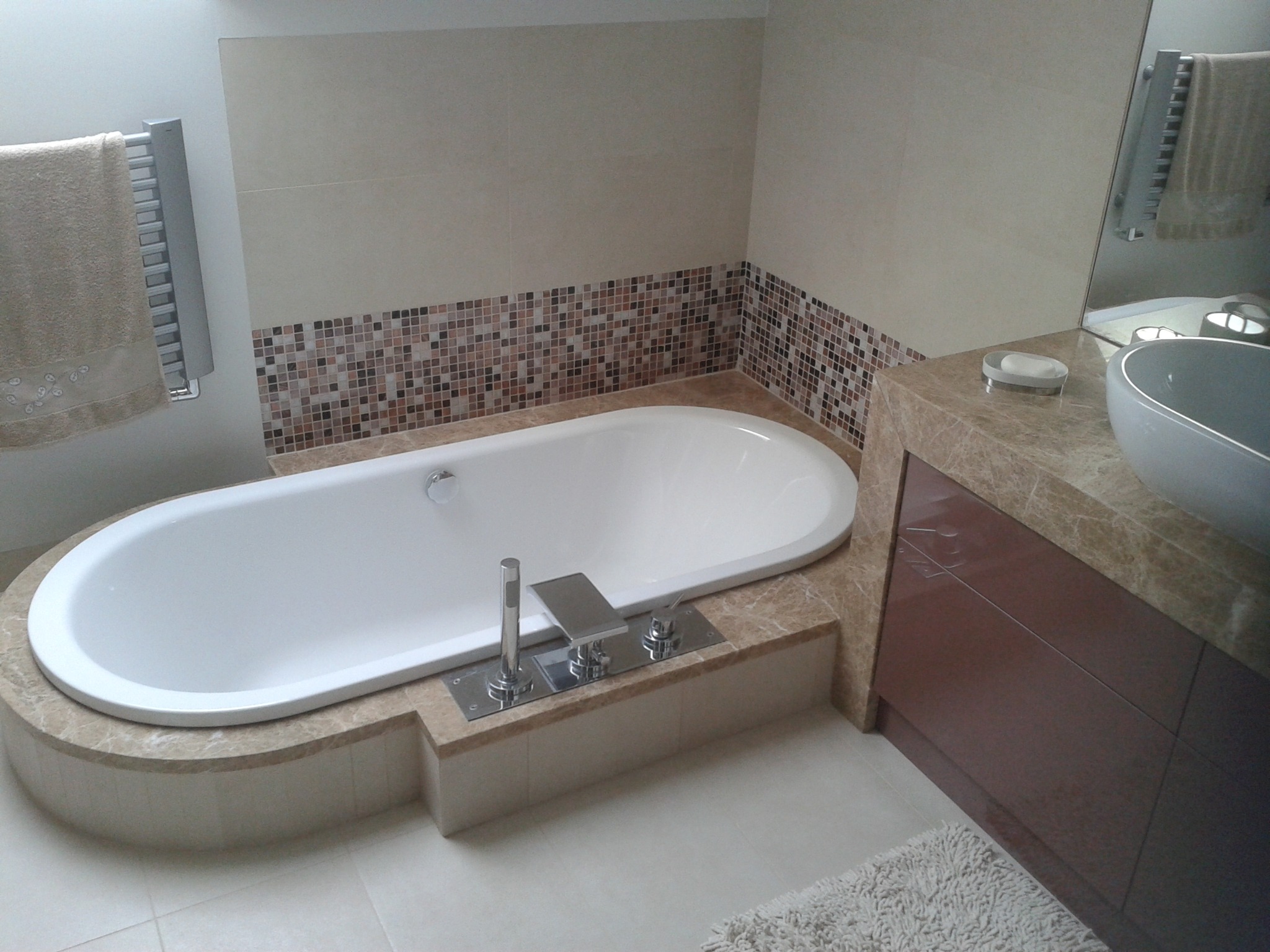 Nowoczesna, owalna wanna w obudowie z beżowego marmuru, bateria wannowa chromowana, ściany wykończone płytkami w odcieniach beżu i brązu, widoczny fragment szafki pod umywalką z blatem z marmuru...