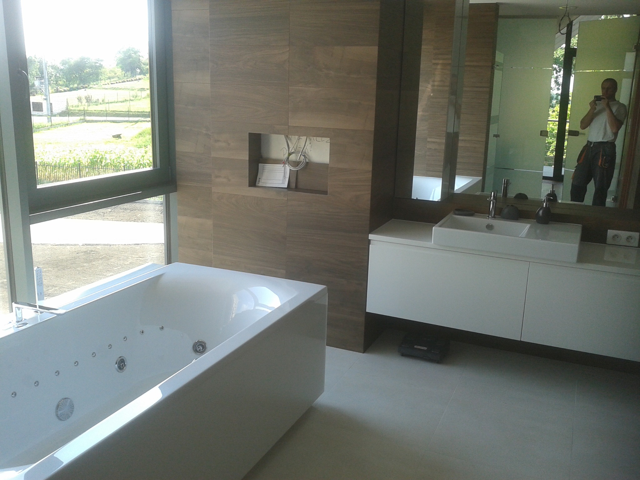 Nowoczesna łazienka z dużą wanną z hydromasażem przy oknie z widokiem na zieleń, drewnopodobne płytki na ścianie z wnęką i białymi kablami, oraz biała szafka z umywalką, odbicie osoby robiącej...