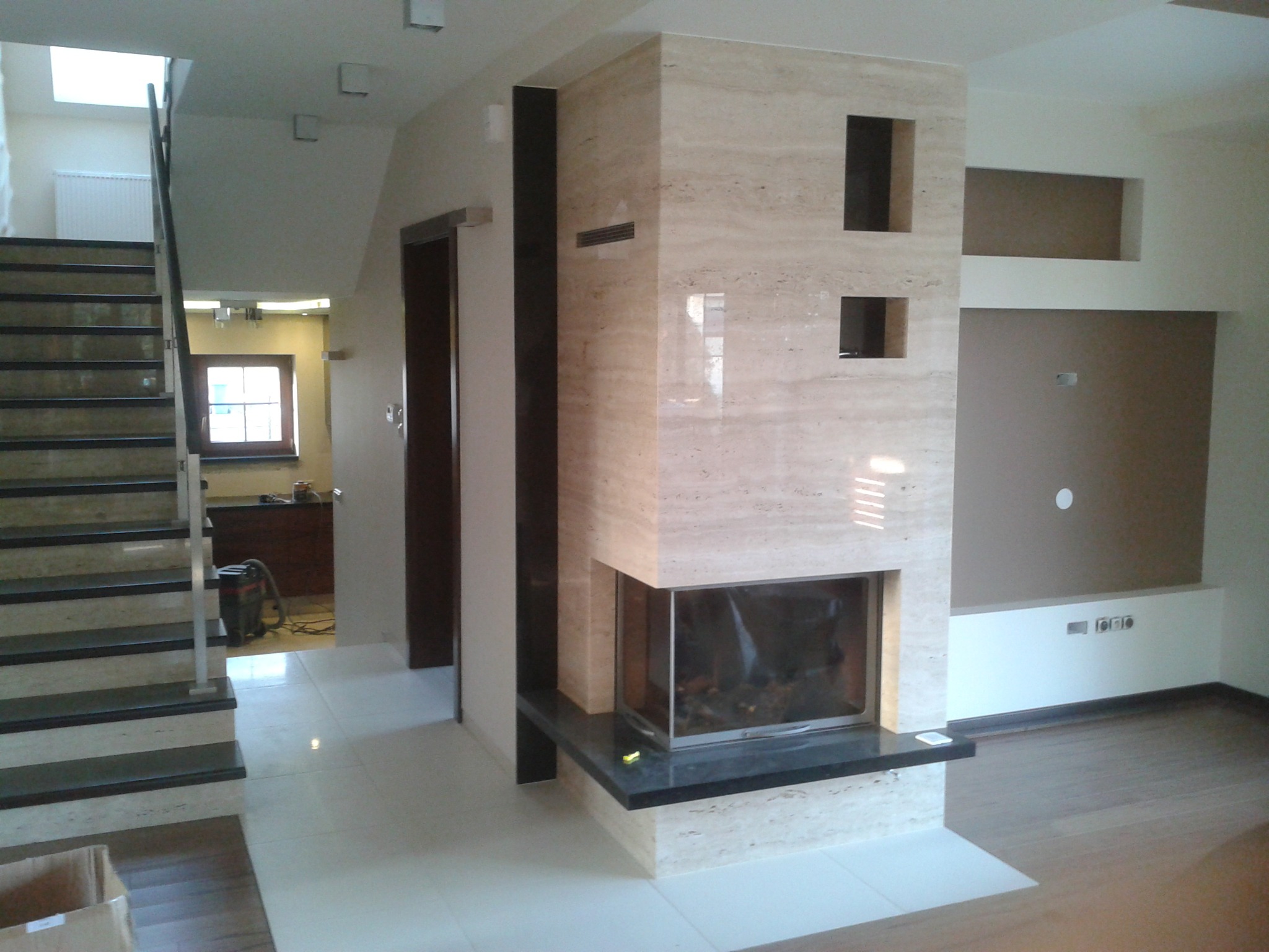 Wykończony salon z marmurowym kominkiem z wkładem, białymi płytkami na podłodze i drewnianą podłogą w tle, widoczne schody z marmurowymi stopniami.