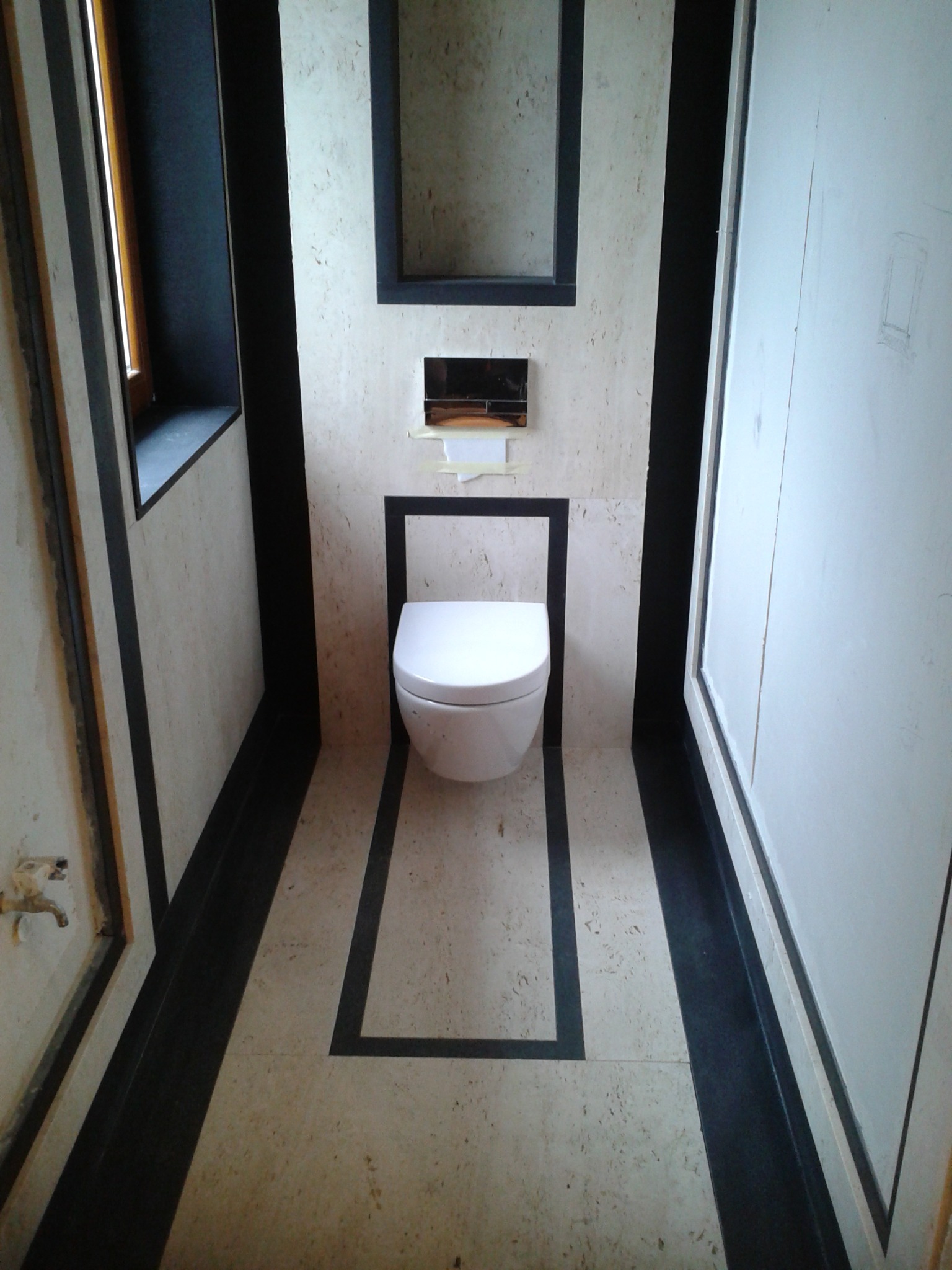 Nowoczesna toaleta w trakcie montażu w łazience z beżowymi płytkami i czarnymi akcentami, widoczne niedociągnięcia wykończeniowe i przyłącze wodne.