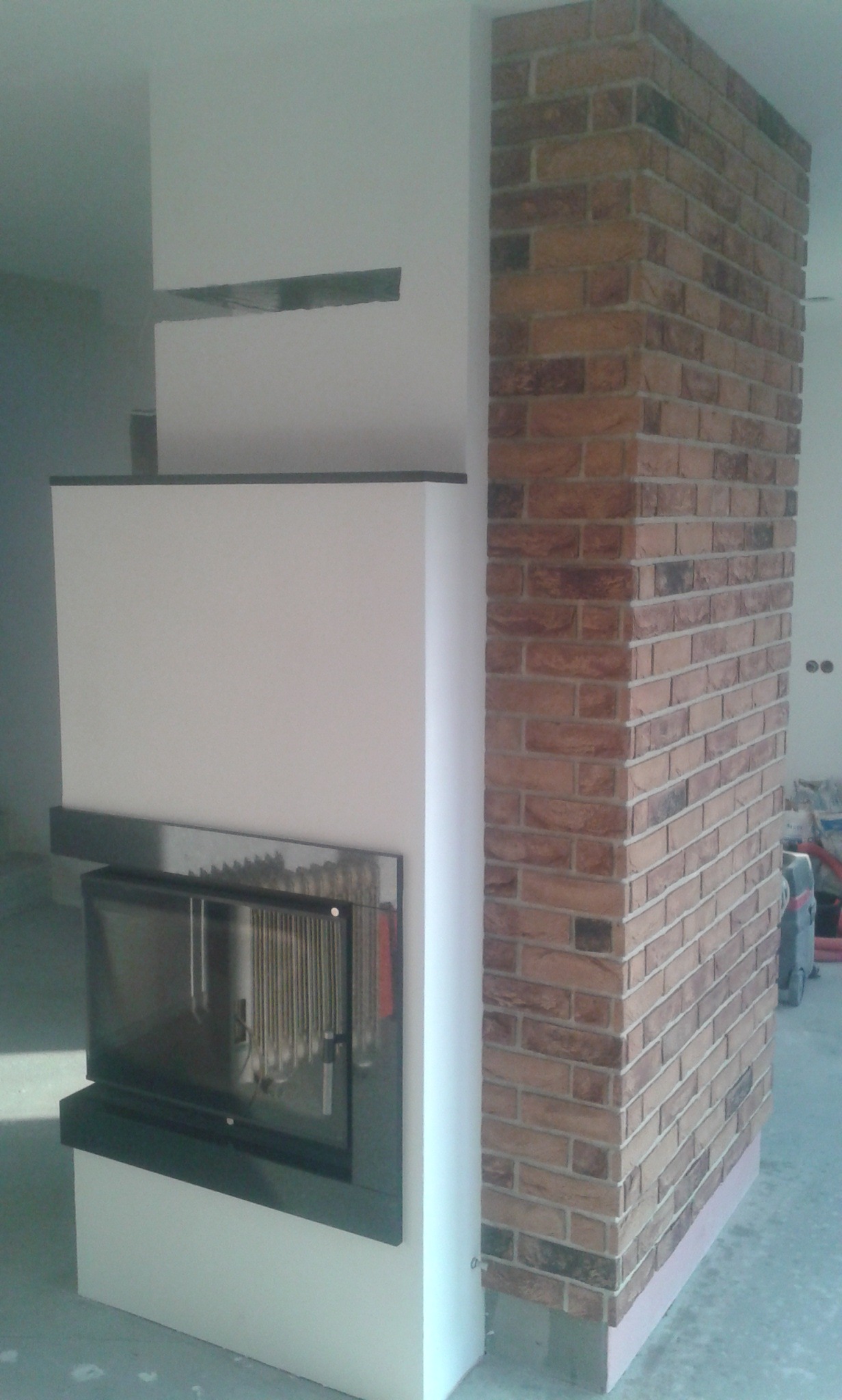 Wnętrze z kominkiem obudowanym białą płytą i ścianą z czerwonej cegły, widoczne przewody instalacyjne w tle.